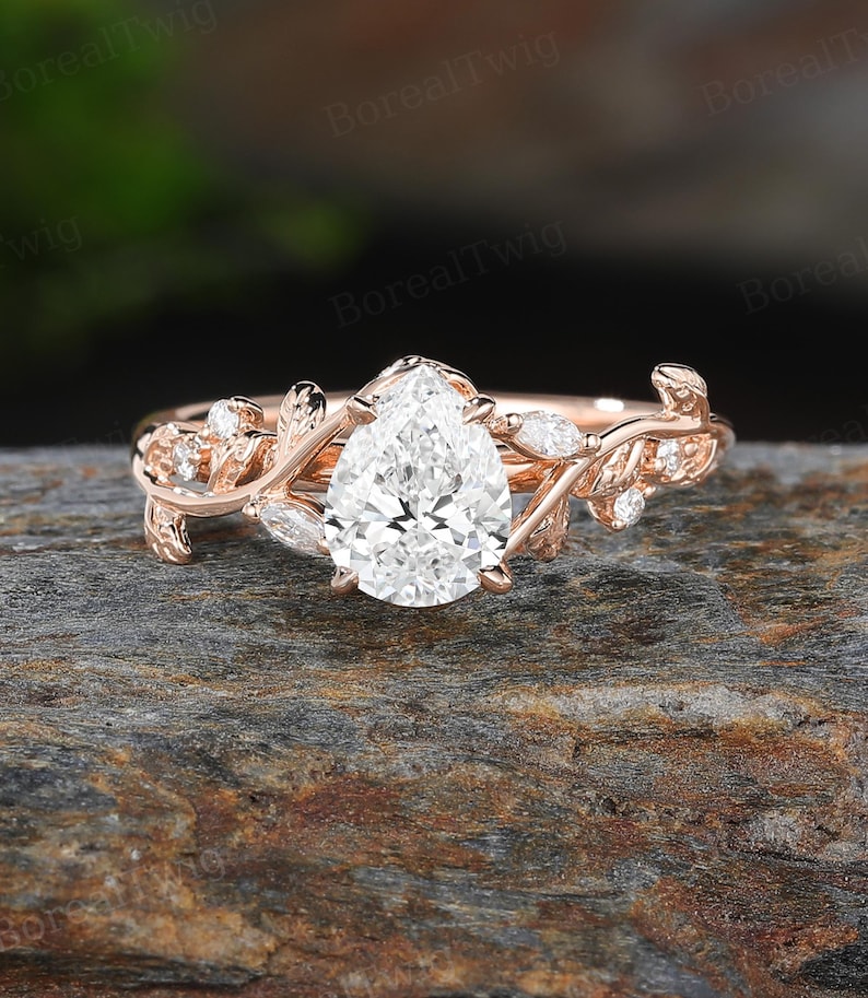 May include: Rose gold engagement ring featuring a pear-shaped diamond center stone. The band is adorned with delicate leaf and vine detailing, and small accent diamonds. The ring is set on a textured, dark gray surface.