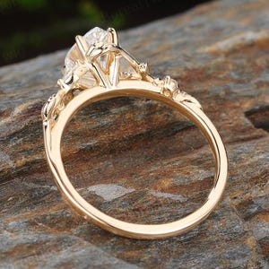 May include: A gold engagement ring with a large, clear gemstone. The ring features intricate floral detailing on the band and around the central stone. The ring is set against a natural, textured background.