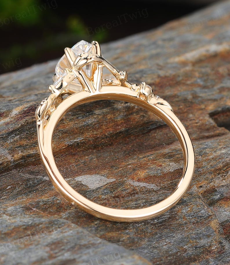 May include: A gold engagement ring with a large, clear gemstone. The ring features intricate floral detailing on the band and around the central stone. The ring is set against a natural, textured background.