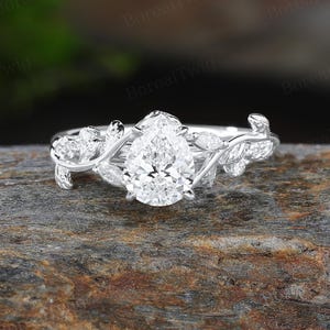 May include: A silver engagement ring with a pear-shaped diamond center stone. The ring features a delicate vine and leaf design on the band, with smaller accent stones. The ring is set on a textured, brown surface.