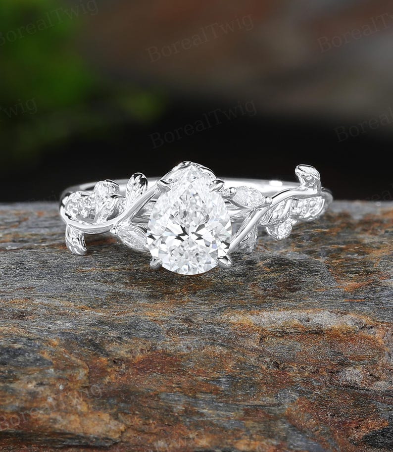 May include: A silver engagement ring with a pear-shaped diamond center stone. The ring features a delicate vine and leaf design on the band, with smaller accent stones. The ring is set on a textured, brown surface.