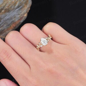 May include: A gold ring featuring a pear-shaped diamond center stone. The band is adorned with delicate leaf and small diamond accents. The ring is shown on a hand, showcasing its intricate design and sparkle. A beautiful piece of jewelry.