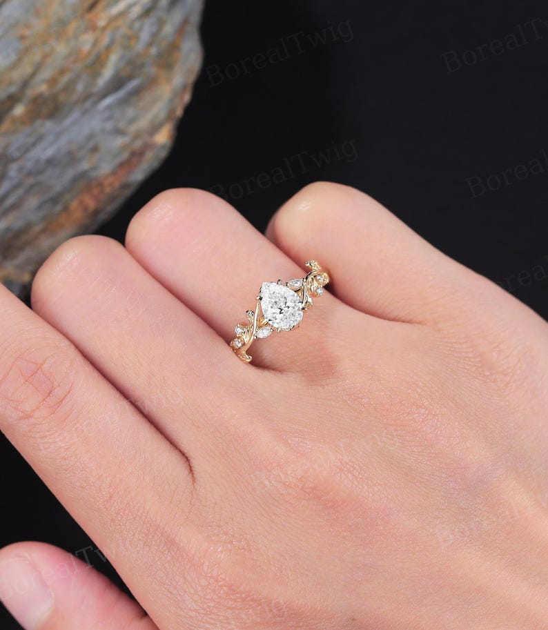 May include: A gold ring featuring a pear-shaped diamond center stone. The band is adorned with delicate leaf and small diamond accents. The ring is shown on a hand, showcasing its intricate design and sparkle. A beautiful piece of jewelry.