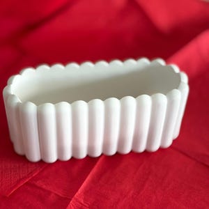 May include: A white, rectangular ceramic planter with a scalloped edge design. The planter is set against a red background, showcasing its clean lines and minimalist aesthetic. Ideal for small plants or succulents.