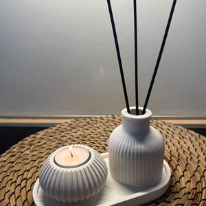Könnte beinhalten: Ein weißer Keramik-Kerzenhalter mit einer brennenden Kerze und eine weiße Vase mit schwarzen Schilf, präsentiert auf einem weißen Tablett. Der Kerzenhalter und die Vase haben ein geripptes Design. Die Artikel befinden sich auf einer gewebten Matte.