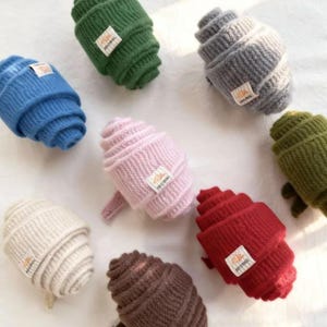 May include: A collection of colorful, rolled knit hats in various shades including blue, green, gray, pink, red, brown, and cream. Each hat features a small rectangular tag with text. The hats are arranged on a white surface.