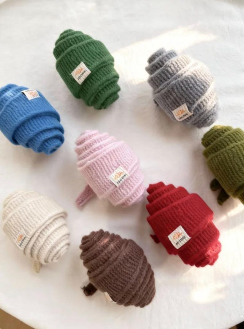 May include: A collection of colorful, rolled knit hats in various shades including blue, green, gray, pink, red, brown, and cream. Each hat features a small rectangular tag with text. The hats are arranged on a white surface.