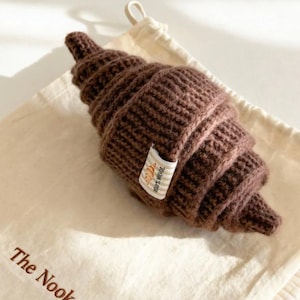May include: A brown, knitted croissant toy with a tag that reads "100% Wool" and a small illustration of a croissant. The toy rests on a cream-colored drawstring bag with the text "The Nook Gift".
