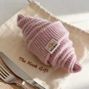 May include: A knitted pink croissant with a small tag that reads "100% Wool". The croissant is resting on a cream-colored cloth bag with the text "The Nook Gift". A fork and knife are placed next to the bag.
