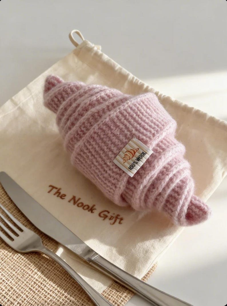 May include: A knitted pink croissant with a small tag that reads "100% Wool". The croissant is resting on a cream-colored cloth bag with the text "The Nook Gift". A fork and knife are placed next to the bag.