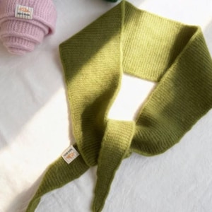 May include: A green knitted scarf with a tag attached, lying on a white surface. Rolled-up knitted hats in pink, blue, and green are visible in the background. The scarf is a solid, muted green color.