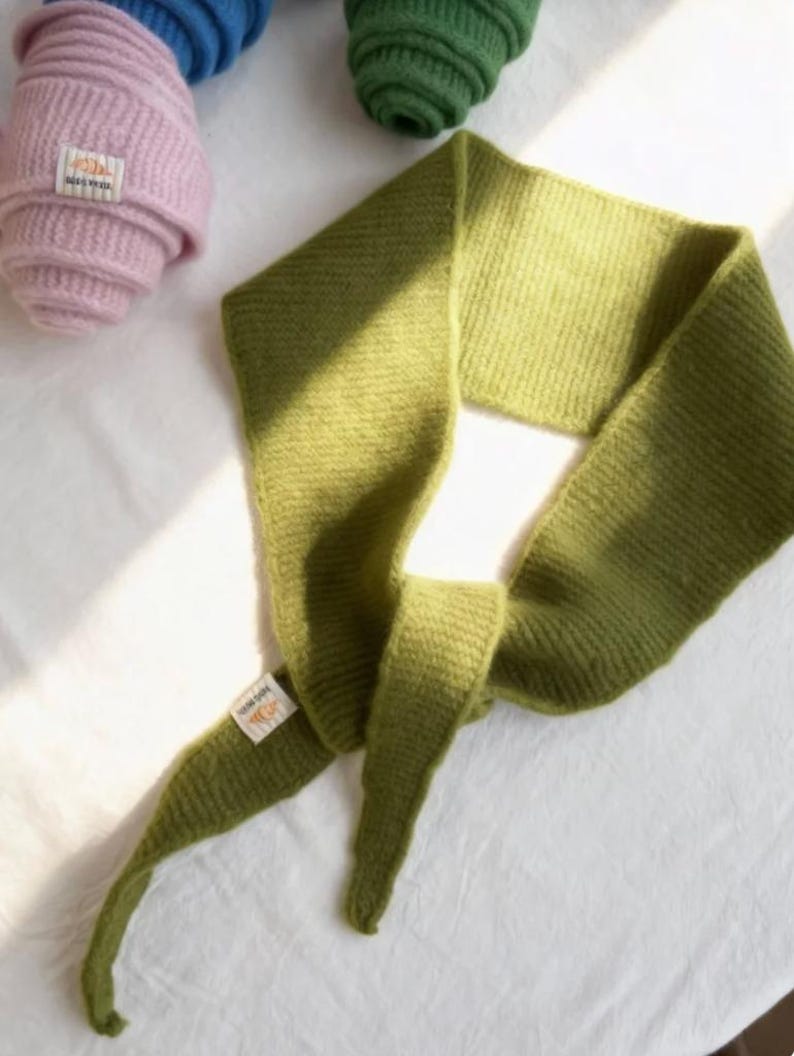 May include: A green knitted scarf with a tag attached, lying on a white surface. Rolled-up knitted hats in pink, blue, and green are visible in the background. The scarf is a solid, muted green color.