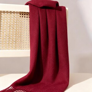 Puede incluir: Una bufanda burdeos con borde de flecos está drapeada sobre el respaldo de una silla blanca. La bufanda está hecha de un material suave y tiene una apariencia texturizada. La silla tiene un respaldo de mimbre.