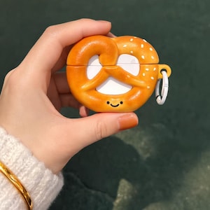 Puede incluir: Una funda naranja con forma de pretzel para auriculares inalámbricos. La funda tiene un interior blanco, una pequeña cara sonriente y detalles de semillas de sésamo blancas. Incluye un pequeño anillo metálico blanco para sujetar.