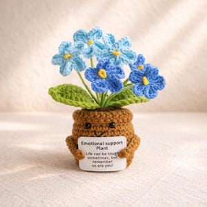 Puede incluir: Una planta de ganchillo con flores azules y hojas verdes en una maceta marrón. La maceta tiene una cara sonriente y sostiene un letrero que dice "Planta de apoyo emocional. La vida puede ser dura a veces, ¡pero recuerda que tú también lo eres!"