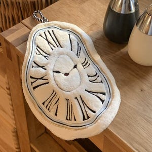 May include: A white, plush clock-shaped decorative item with black Roman numerals and hands. The clock face has a melting appearance, with a black and white striped loop for hanging. The item is on a wooden surface.
