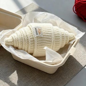 May include: A cream-colored, crocheted croissant rests in a beige container lined with white paper. A small label with text and a croissant graphic is attached. A ball of red yarn is visible in the background.