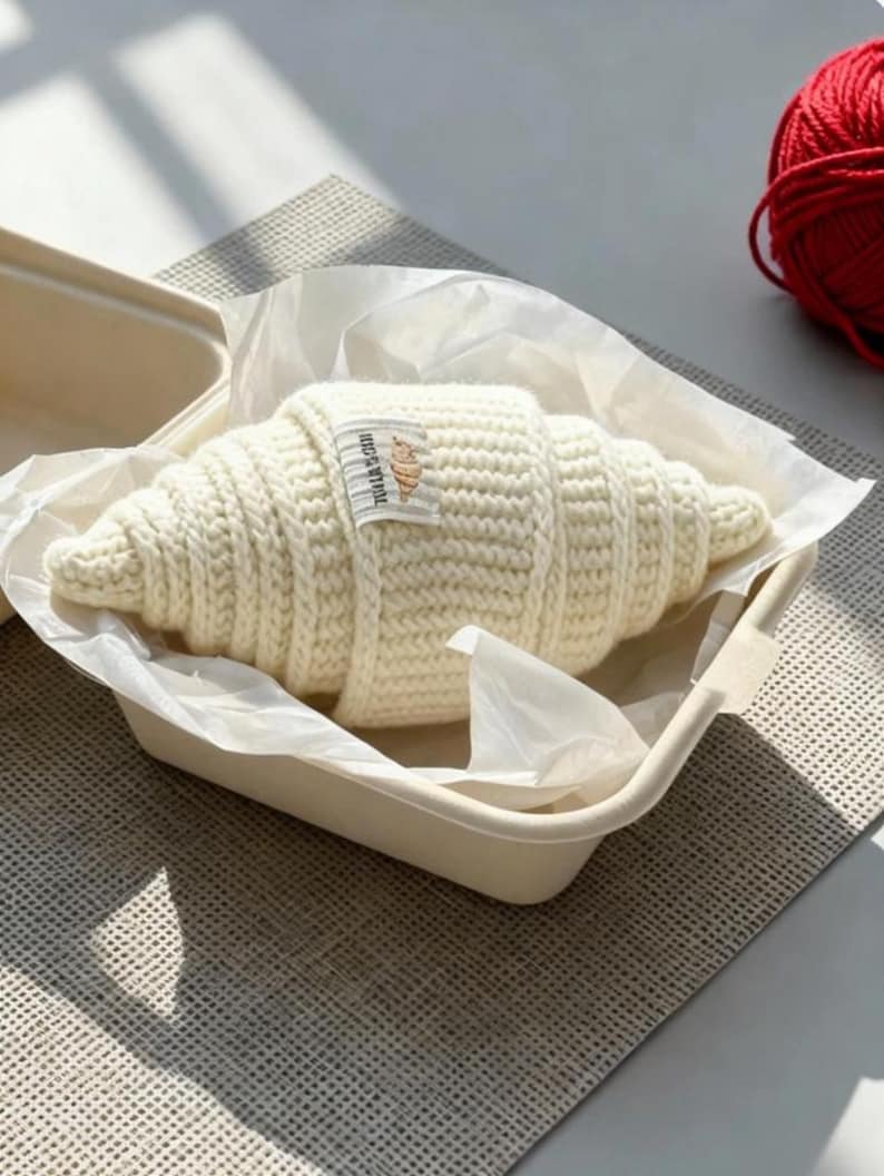 May include: A cream-colored, crocheted croissant rests in a beige container lined with white paper. A small label with text and a croissant graphic is attached. A ball of red yarn is visible in the background.