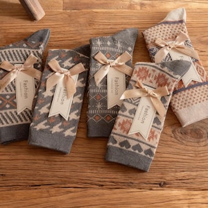 Op de afbeelding: Vijf paar sokken met patronen en "Fashion" labels liggen op hout. De sokken hebben geometrische en hartpatronen in grijs, bruin en crème, vastgebonden met beige linten. De sokken zijn ongeveer 25 cm lang.