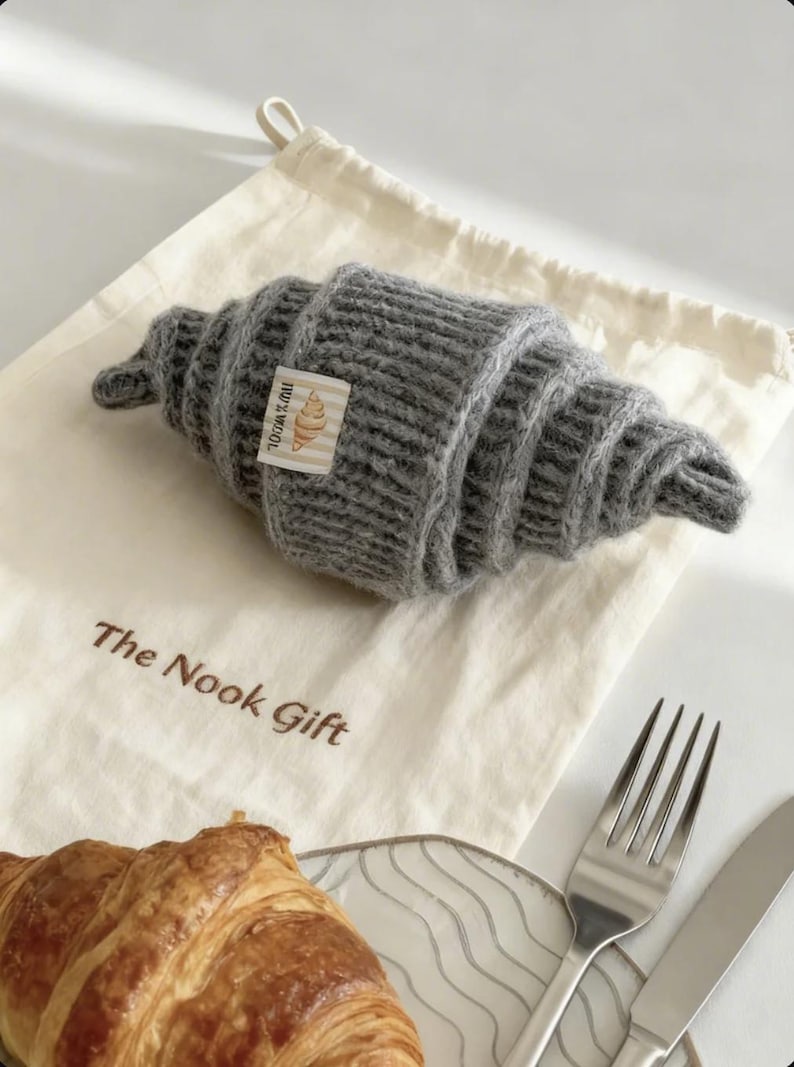 以下が含まれることがあります： グレーのニットクロワッサンが、「The Nook Gift」の文字が入ったクリーム色の巾着袋の上に置かれています。クロワッサンには「100% Wool」と書かれたタグが付いています。本物のクロワッサン、フォーク、ナイフも写っています。