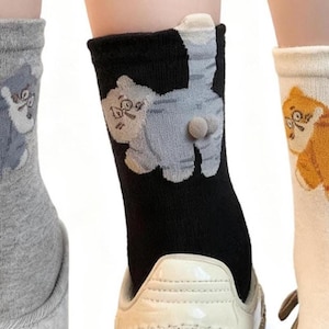 May include: Three pairs of novelty socks, each featuring a cartoon cat design on the back. One pair is gray, another black, and the third is white with orange stripes. The socks have a 3D element for the cat's rear.