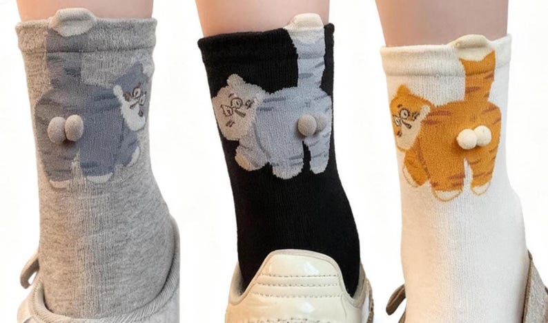 May include: Three pairs of novelty socks, each featuring a cartoon cat design on the back. One pair is gray, another black, and the third is white with orange stripes. The socks have a 3D element for the cat's rear.