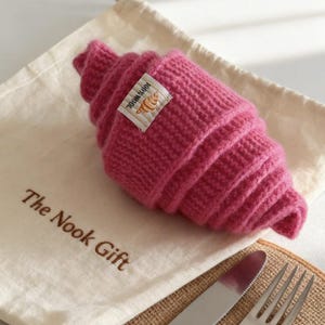 May include: A knitted, hot pink croissant rests on a cream-colored bag with the text "The Nook Gift". A small tag on the croissant reads "100% Wool". A knife and fork are visible in the foreground.