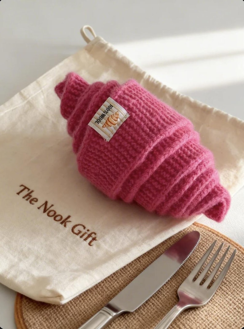 以下が含まれることがあります： 「The Nook Gift」の文字が入った白い巾着袋の上に、編み物のピンク色のクロワッサンが置かれています。「100% Wool」とクロワッサンのイラストが入った小さなタグ。ナイフとフォークが見えます。