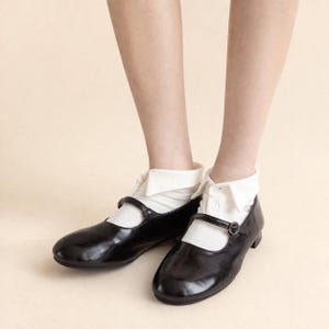 May include: Black leather Mary Jane shoes with a strap and buckle. The shoes are worn with white socks and a white collared shirt. The shoes have a rounded toe and a low heel.