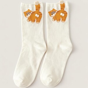 May include: A pair of white socks featuring an embroidered orange tabby cat design. The cat is depicted from the rear, with its striped tail raised. The socks are made of a soft, comfortable material.