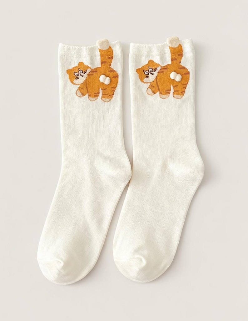 May include: A pair of white socks featuring an embroidered orange tabby cat design. The cat is depicted from the rear, with its striped tail raised. The socks are made of a soft, comfortable material.