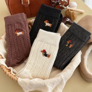 May include: A selection of cable-knit socks in shades of brown, black, cream, and grey. Each sock is adorned with an embroidered dachshund wearing an outfit. The socks are presented in a woven basket.