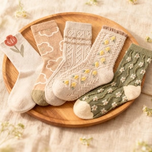 May include: Five pairs of cozy socks in various colors and patterns are displayed on a wooden tray. Designs include a tulip, clouds, cable knit, yellow flowers, and white flowers on a green background. The socks are made of a soft, textured material.