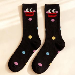 以下が含まれることがあります： 漫画の顔のデザインとカラフルな水玉模様が特徴の黒い靴下のペア。顔には赤い唇、白い目、文字「CC」があります。水玉模様はピンク、青、黄色です。楽しくて個性的なアクセサリーです。