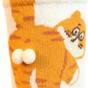 May include: White and orange socks with a cartoon tiger design. The tiger has orange stripes and a smiling face. Two white pom-poms are attached to the back of the sock. The socks are made of a soft, knitted material.