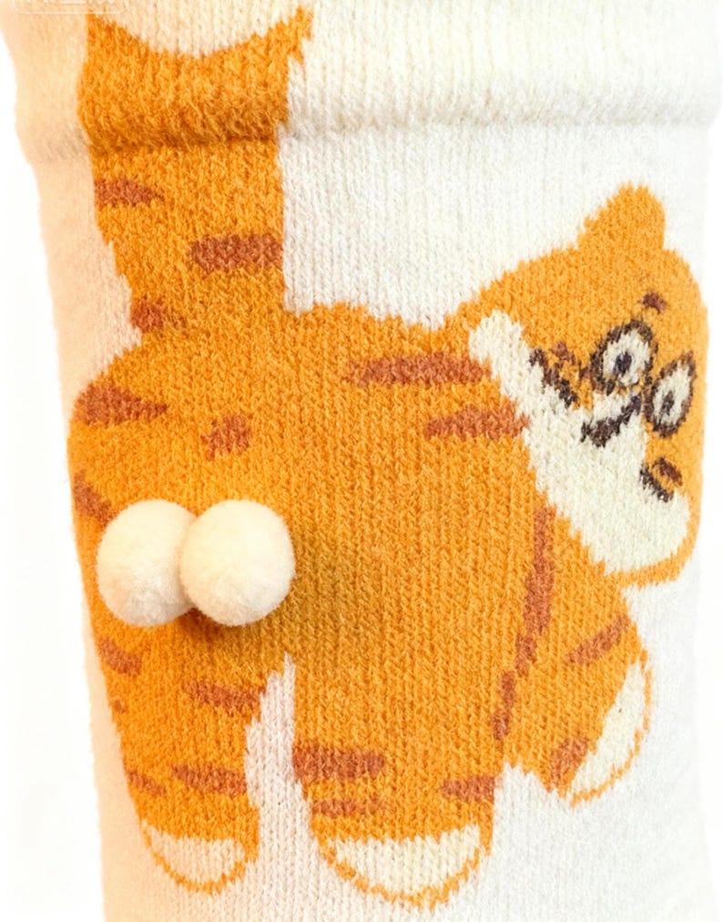 May include: White and orange socks with a cartoon tiger design. The tiger has orange stripes and a smiling face. Two white pom-poms are attached to the back of the sock. The socks are made of a soft, knitted material.