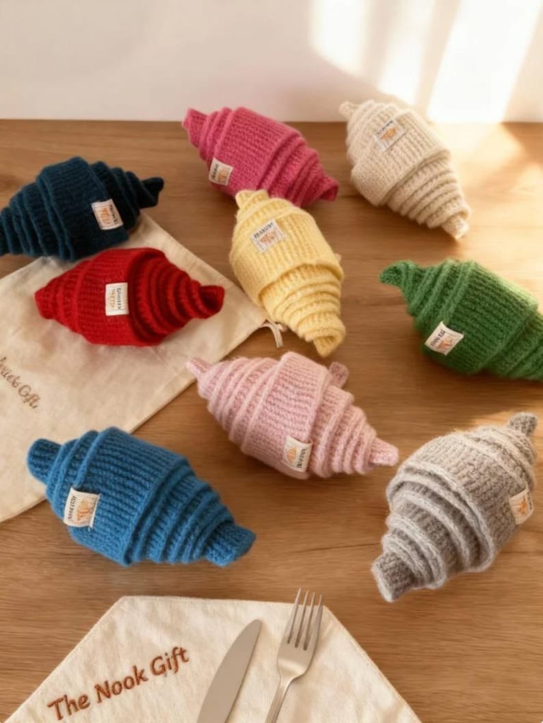 May include: A collection of colorful, knitted croissant-shaped items. The croissants are in various colors, including blue, pink, red, yellow, green, and gray. Each croissant has a small tag. The items are arranged on a wooden surface with a napkin, knife, and fork.