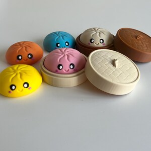 May include: A collection of small, round, colourful silicone-like dumpling figures with smiling faces, including yellow, orange, blue, and pink versions, some resting inside miniature circular bamboo-style steamer baskets with lids.