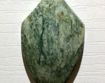 Puede incluir: Una gema en forma de escudo con un patrón verde moteado. La piedra tiene una parte superior puntiaguda y una parte inferior redondeada, con variaciones de color de verde claro a oscuro. La superficie tiene un aspecto natural y texturizado.