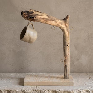 Peut inclure: Un porte-tasse en branche de bois naturel avec une base rectangulaire en bois clair et plusieurs crochets en métal couleur laiton. Une tasse en céramique beige de forme arrondie est suspendue à l'un des crochets de la branche horizontale.