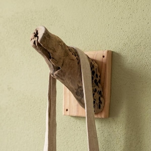 May include: A wall-mounted hook featuring a piece of weathered, textured driftwood attached to a square, light-coloured wooden base. A long, beige fabric strap hangs over the driftwood branch, which displays natural holes and a rough, organic surface.