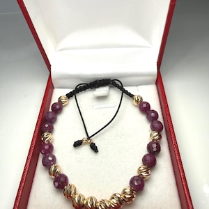 Puede incluir: Una pulsera de cuentas con cuentas doradas texturizadas alternas y cuentas facetadas de color rojo intenso. La pulsera se muestra dentro de una caja de joyería roja con interior blanco. Tiene un cordón negro ajustable.