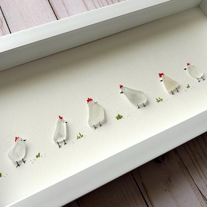 Puede incluir: Una obra de arte enmarcada que presenta una l&iacute;nea de pollos blancos hechos de vidrio de mar con crestas rojas. Los pollos est&aacute;n dispuestos en una fila sobre un fondo blanco con hierba verde pintada debajo.