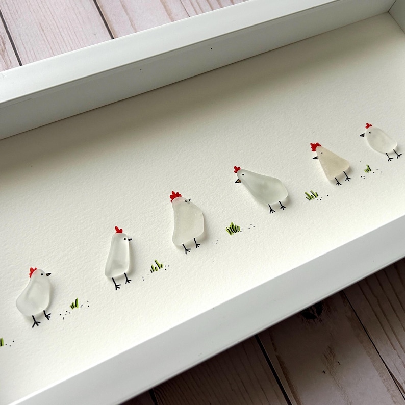Puede incluir: Una obra de arte enmarcada que presenta una l&iacute;nea de pollos blancos hechos de vidrio de mar con crestas rojas. Los pollos est&aacute;n dispuestos en una fila sobre un fondo blanco con hierba verde pintada debajo.