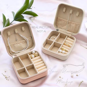May include: Two open, square jewellery boxes in pink and cream, displaying various jewellery pieces. The pink box holds a pearl necklace and earrings, while the cream box displays rings and necklaces. The text "REAL PICTURE NOT AI" is visible.
