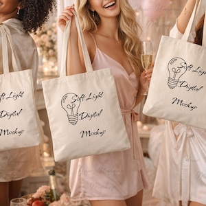 May include: Three off-white tote bags with the text "Soft Light Digital Mockup" and a lightbulb graphic in black. The bags are held by three people, each wearing a robe and holding a glass of champagne.