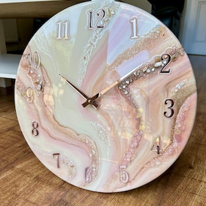 Può includere: Un orologio da parete rotondo con un design marmorizzato nei toni del rosa, crema e oro. Il quadrante presenta numeri e lancette metalliche in rilievo. L'orologio ha un diametro di circa 30 cm.