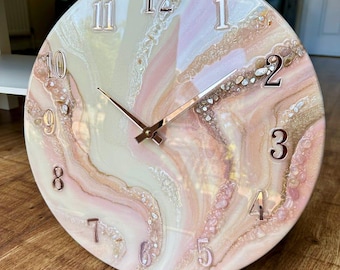 Puede incluir: Un reloj de pared redondo con un diseño veteado en tonos rosa, crema y dorado. La esfera del reloj presenta números y manecillas metálicas en relieve. El reloj tiene aproximadamente 30 cm de diámetro.