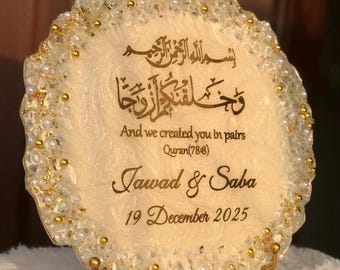May include: A decorative, round, cream-colored resin piece with gold accents and embedded pearls. The piece features Arabic calligraphy and the English text "And we created you in pairs," along with names and a date.