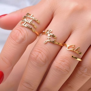 Peut inclure: Plusieurs bagues dorées avec les noms Fabrice, Pauline, Cérine, Rose, Jude et Celia écrits en cursive. Les bagues sont portées sur des doigts vernis de rouge. Le nom de la marque "AnissaSouk" est visible.
